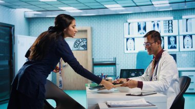 Erkek doktor röntgen görüntüleme ve laboratuvar sonuçlarını değerlendiriyor, hasta bir hastaya rapor vermek için hastane kayıtlarına bakıyor. Sağlık uzmanları sigorta için iyileşme planı hazırlıyor. Kamera B.