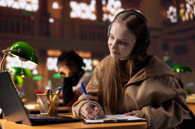 Genç öğrenci, Webinar 'la video konferansı yoluyla öğretmenden notlar alıyor, kampüs kütüphanesinden online derse katılıyor. Üniversite öğrencisi akademik web konferansından bilgi yazar.