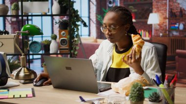 Evde klavyede yazı yazarken abur cubur yemekle meşgul. İnternetteki iş görevlerini çözmek için elektronik cihaz kullanırken kruvasan seven kadın, kamera A