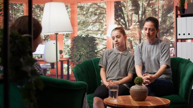 Mother and teenage daughter engaging in a confidential therapy session, discussing with the adolescent and mom about her emotional issues and mental health with a trustworthy therapist. Camera B. clipart