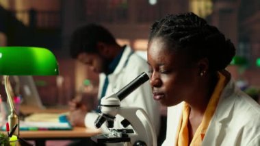 Dişi bilim öğrencisi, kampüs kütüphanesindeki bilimsel araştırmaları için mikroskop altında hücreleri inceliyor, kimyasal süreçler ve teziyle ilgili notlar alıyor. Kamera B.