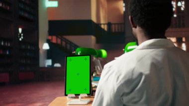 Genç tıp öğrencisi kroma anahtarının yanındaki sınavlar için araştırma yapıyor, yeşil bir lamba altında farmakoloji belgelerini gözden geçiriyor. Sınav hazırlıklarına ve tıp kariyerlerine odaklanıyorlar. Kamera A.