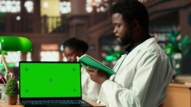 Tıp öğrencisi yeşil ekranın yanındaki kampüs çalışma alanında araştırma, anatomi dersleri ve akademik yöntemleri birleştiren kitaplar kullanıyor. Öğrenci tıp pratiği için araştırma kağıtları okuyor. Kamera B.