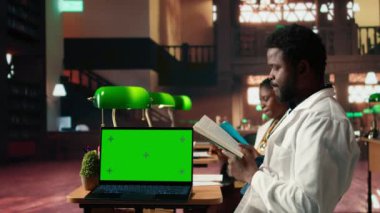 Afro-Amerikalı tıp öğrencisi fotokopi alanının yanında sağlık hizmeti kitabı okuyor, klasik bir kütüphanede yeşil lambalar altında tıbbi uygulama yöntemlerine odaklanıyor. Eğitime bağlılık. Kamera A.