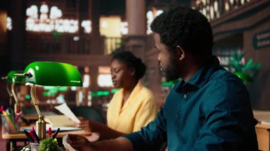 Siyahi üniversite öğrencileri üniversite kütüphanesinde sınıf notlarını karşılaştırıyor, akademisyenler fotokopi kitaplarından bilgi alıyor ve danışmanlık yapıyor. Öğrenciler yaklaşan sınavlarına hazırlanıyor. Kamera A.
