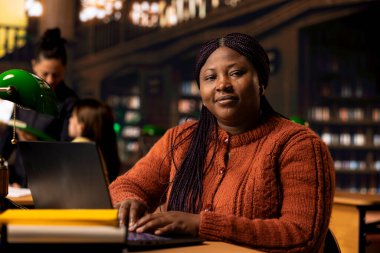 Sessiz bir kütüphane çalışma alanında dizüstü bilgisayarında yazı yazan siyahi özgüvenli kütüphanecinin portresi, kaynakları organize etmeye ve öğrencilere akademik yolculuklarında gurur ve uzmanlıkla rehberlik etmeye adanmıştır..