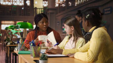 Afrikalı Amerikalı akıl hocası, kız öğrencilerine destek veriyor. Kütüphanede ders programı üzerinden ders veriyor. Profesör öğrenme malzemeleri sağlıyor, çocuklara akıl hocalığı yapıyor. Kamera A.