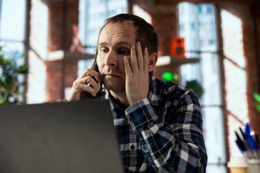 Man seated at home office desk answering perturbing phone call informing him of bad news while checking emails on laptop. Worker alarmed by distressing telephone call with friend at work clipart