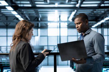 Veri merkezindeki bilgisayar bilimcileri dizüstü bilgisayar ve tablet kullanıyorlar, güvenlik yamalarını çalıştırıyorlar, güvenlik duvarı yapılandırmalarını doğruluyorlar. Kadın ve erkek sunucu çiftliğinde ekipman kırılganlığı taraması yapıyor.