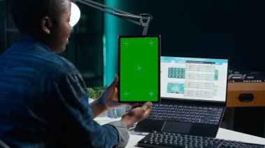 Çalışırken elinde yeşil ekran tableti olan bir kadın, evdeki ofis masasında oturuyor. Evdeki kişi dizüstü bilgisayardaki borsa grafiklerine ve model cihazına bakıyor.
