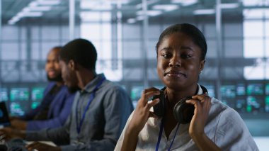 Portrait of smiling engineer hearing podcasts to make time pass faster while working in server farm to optimize it for data processing requirements. Happy woman listens songs in data center, camera A clipart