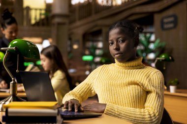 Afrikalı Amerikalı liseli kız kütüphanedeki bir çalışma masasında ders notları yazıyor, bir okul projesi üzerinde bilgisayarında online bilgi ile sıkı çalışıyor. Öğrenci akademik enstitüde ders için hazırlanıyor.