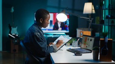 Evde çalışan, dizüstü bilgisayar kullanan, istatistik tablolarına bakan Afrikalı Amerikalı bir kadın. Kişisel apartman ofisindeki uzaktan çalışan, not defteri ve not defteri arasındaki önemli bilgileri karşılaştıran, kamera A