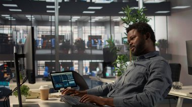 Ofiste kollarını ve sırt kaslarını esneten Afrikalı Amerikalı bir çalışan idari bir görevi bitirdikten sonra sandalyesinde dinleniyor. Erkek analist şirket için hedefler belirliyor. Kamera B.