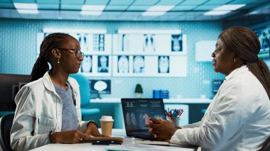 Siyahi pratisyen hekim ve hasta bir klinikte sağlık kontrolü ve tedavi seçeneklerini tartışıyor, iletişim ve tıbbi uzmanlığı vurguluyor. Kadın uzman tavsiye veriyor. Kamera B.