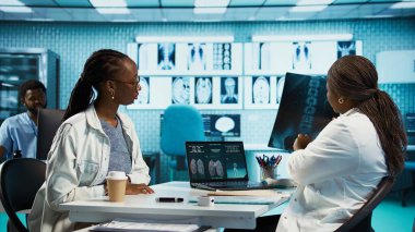 Siyahi hasta, tıbbi kabinedeki kadın doktorla görüşüyor. Kişisel bakım için MRI ve röntgen analizini tartışıyorlar. Sıhhiye, tarama sırasında sağlık ve rehberlik sunar. Kamera B.