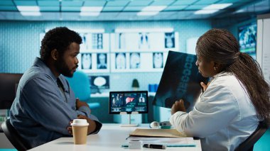 Siyahi erkek hasta, tarama için sıhhiyeye röntgen testi sonuçlarını getiriyor. Teşhis raporlarını inceliyor. Tıbbi uzman kabinedeki radyografi filmini değerlendiriyor. Kamera B.