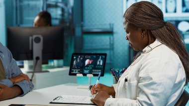 Afrika kökenli Amerikalı doktor ve hasta tıbbi sonuçlar hakkında derinlemesine konuşuyorlar, sağlık ve tedavi planı oluşturmak için sağlık raporlarını kullanıyorlar. Önerilen ilaç listesi. Kamera A.