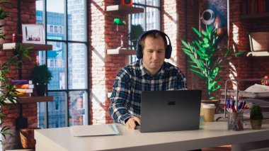 Teleworker şirket projesi için verileri araştırırken müziğin tadını çıkarıyor, kişisel ev ofisinde görevler yapıyor. Adam dizüstü bilgisayarın önünde oturuyor ve kulaklık ve kamera A kullanıyor.