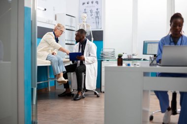 Siyahi doktor, tedavi planını özetlemek için pano kullanarak emekli beyaz kadınla sohbet ediyor. Kafkasyalı yaşlı hasta, hekim hastane odasında dikkatle dinlerken sorular soruyor..