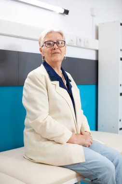 Yaşlı beyaz kadın klinikte otururken kameraya bakar, tıbbi randevu için hazırlanır. Hastane yatağında oturmuş, sakince sağlık kontrolü için doktorunu bekleyen yaşlı kadının portresi..