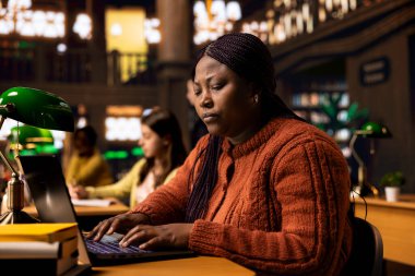 Sessiz bir kütüphane çalışma alanında dizüstü bilgisayarında yazı yazan mutlu kadın kütüphaneci, kendini kaynakları organize etmeye ve öğrencilere akademik yolculuklarında gurur ve uzmanlıkla rehberlik etmeye adadı..