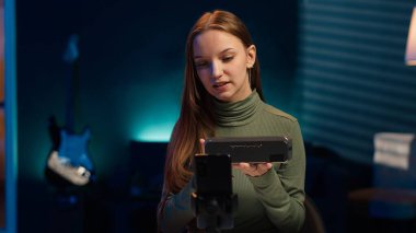 Etkileyici, taşınabilir hoparlör performansını değerlendiriyor, kullanılan parçalardan bahsediyor, telefonla çekim yapıyor. Tripodun üzerinde akıllı telefon kullanan kız müzik cihazı incelemesi, kullanılmış malzemelerden memnun, kamera A