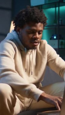 Dikey Video Siyah uzaktan kumandalı işçi, neon ışıklarla dekore edilmiş şık dairesinde dizüstü bilgisayarında gezinirken esnekliğin ve konforun tadını çıkarıyor. Rahat bir ortamda mükemmel bir şekilde yakalanmış serbest çalışma.