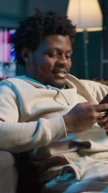 Dikey Videolu Serbest Çalışan Cep telefonuyla eğlenen yakın bir bilgisayarla akıllı telefonundan arkadaşlarına mesaj atan biri. Konforlu oturma odası vitrinleri ve uzak yaşam tarzında eğlence.