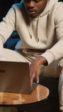 Dikey Video Odaklı Adam evde bir iş projesi için çevrimiçi araştırma yapıyor, kahve masasında dizüstü bilgisayarla oturuyor ve internette makaleler okuyor. Web sayfalarında gezinen ve toplanan kişi