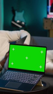 Dikey Video Siyah uzaktan kumandalı işçi, Chroma Key 'in yanındaki koltukta boş zamanını geçiriyor, kulaklıklı bir podcast izlemek için tabletini kullanıyor. Genç, serbest çalışan, kendi evinde rahatlıyor.