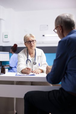 Emekli kadın doktor hastane konsültasyonu sırasında yaşlı erkek hastalara sağlık bilgileri veriyor. Emekli kadın doktor, yaşlı adamla sağlık tedavisi hakkında konuşuyor..