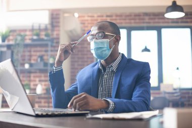 Düşünceli siyah işadamı dizüstü bilgisayar kullanıyor, alnına tuğla duvardaki kalemle vuruyor. Maskeli bir yönetici. Masada yeni bir iş araştırması yapmaya odaklanmış..