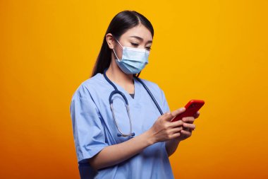 Tıbbi üniformalı ve yüz maskeli genç hemşire izole edilmiş arka planda dikilirken cep telefonuyla mesaj atıyor. Steteskoplu Asyalı kadın doktor, cep telefonu kullanıyor, sosyal medya hesaplarını kontrol ediyor..