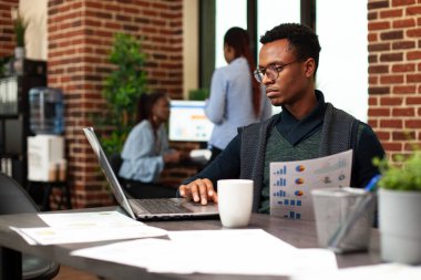 Elinde evrak tutan Afrikalı Amerikalı girişimci cihaz ekranına bakıyor, proje planlarını hazırlıyor. Erkek çalışan, elinde belge ve dizüstü bilgisayarla masada oturmuş şirket verilerini araştırıyor..