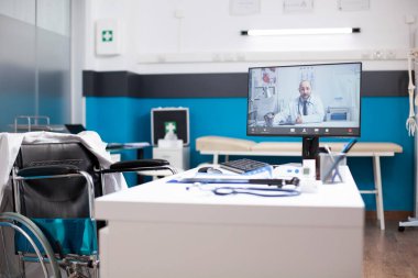 Vacant clinic room with desktop pc showing male physician conducting online consultation. Empty medical office for doctor in wheelchair shows growing role of technology in remote healthcare services. clipart