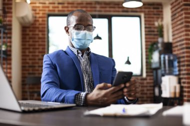 Tuğla duvardaki ofisinde cep telefonunda yüz maskesi olan bir iş adamının portresi. Afrikalı Amerikalı menajer işe ara veriyor, sohbet etmek ve sosyal medyayı kontrol etmek için akıllı telefon kullanıyor..