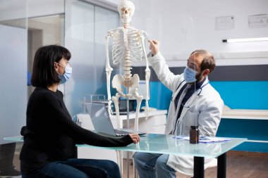 Doktor, tıbbi danışmanlık sırasında hasta bir kadınla iskelet yapısını inceliyor. Erkek kayropraktik uzmanı, masadaki kadın hastaya kemik tedavisi seçeneklerini açıklıyor. Klinikteki koruyucu ekranla ayrılmış.