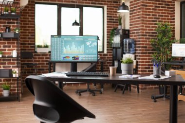 Boş başlangıç ofisi, finansal tabloları gösteren bilgisayar monitörlü modern bir masaya sahip. Masaüstü bilgisayarı, dizinleri, kutuları ve iyi düzenlenmiş tuğla duvar çalışma alanını tamamlayan bitki süslemeleri olan raflar.
