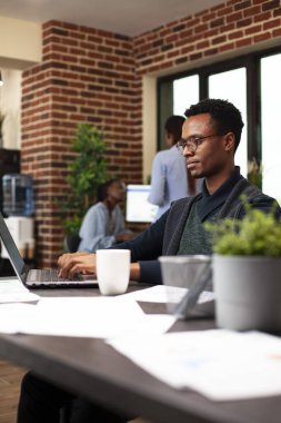 Siyah işadamı masasına oturmuş, dijital cihazını kullanarak iş e-postalarına cevap veriyor ve günlük işleri gözden geçiriyordu. Dizüstü bilgisayarında yazan erkek girişimci, modern ofiste yeni bir iş projesine hazırlanıyor..