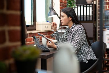 Kadın girişimci, iş analizlerini gösteren bilgisayar ekranına odaklanıyor. Masaüstü monitöründeki bilgilerin karşılaştırılması ve analiz edilmesi için, Asyalı yönetici elinde mali grafiklerle kalem ve pano tutuyor.