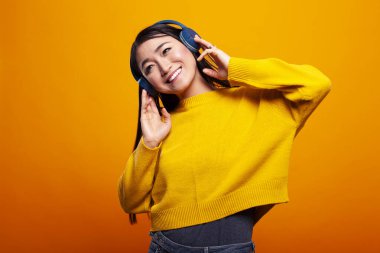 Neşeli bir suratı olan Asyalı bir bayan canlı bir arkaplan karşısında kablosuz kulaklıkla müzik dinlemekten hoşlanıyor. Sarı kazaklı gülümseyen genç kadın en sevdiği sesi dijital bir aletle dinliyor..