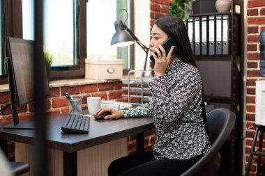 İş kadını masasında oturuyor, cep telefonuyla bir müşteriyle konuşuyor ve bilgisayarındaki güncellemeleri paylaşıyor. Kadın çalışan monitöre bakıyor ve müdürle telefon görüşmesi yapıyor..