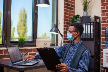 Siyah yönetici gözlüklü masada oturuyor, şirket araştırma belgelerini gözden geçirmeye ve dizüstü bilgisayardaki çizelgelerle karşılaştırmaya odaklanmış. Elinde pano olan bir işadamı, aygıt üzerindeki bilgileri güncelliyor.