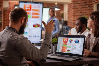 Sakallı adam dizüstü bilgisayardan veri okuyor ve başlangıç ofisinde strateji planlama toplantısında gruba güncelleme sağlıyor. Yükselmiş elli bir erkek çalışan, iş sunumunda siyahi kadın çalışana yardım ediyor.