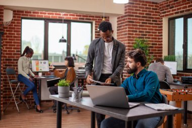 Erkek profesyonel belgeler tutuyor ve müdürüyle fikirlerini paylaşıyor. Tuğla duvar çalışma alanında beyin fırtınası stratejileri uyguluyor. Elinde kağıtları olan Afrikalı Amerikalı analist, sakallı iş arkadaşına geri bildirim veriyor..