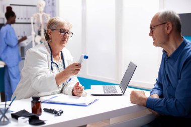 Kıdemli doktor yaşlı adamın sağlığını tıbbi konsültasyonda izlemek için dijital termometre kullanıyor. Emekli emekli hasta, klinikte ateşini kontrol etmek için emekli kadın doktoru bekliyor..