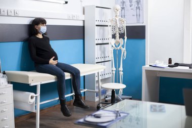 Expectant mother in face mask gently holds her pregnancy belly, awaiting checkup visit in clinic room filled with medical tools. Pregnant woman seated on hospital bed ready for health examination. clipart