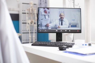 Klinik masası, masaüstü bilgisayarında, sağlık uzmanını gösteren aktif sanal bir oturum düzenliyor. Bilgisayar ekranında beyaz erkek doktorla uzaktan tıbbi konsültasyon sunan bir video çağrısı var..