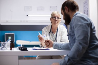 Genç erkek hasta klinikteki yaşlı kadın doktordan yazılı tıbbi talimatlar alıyor. Emekli pratisyen doktor, sakallı adama ilaç için reçeteli kağıt sağlıyor..